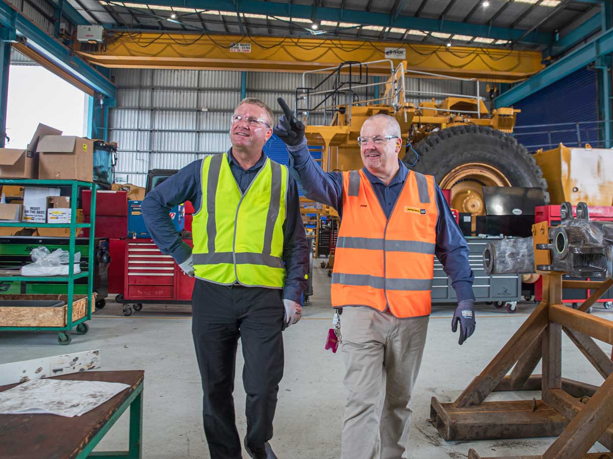 Two men point as they walk through a workshop containing mining vehicles and equipment