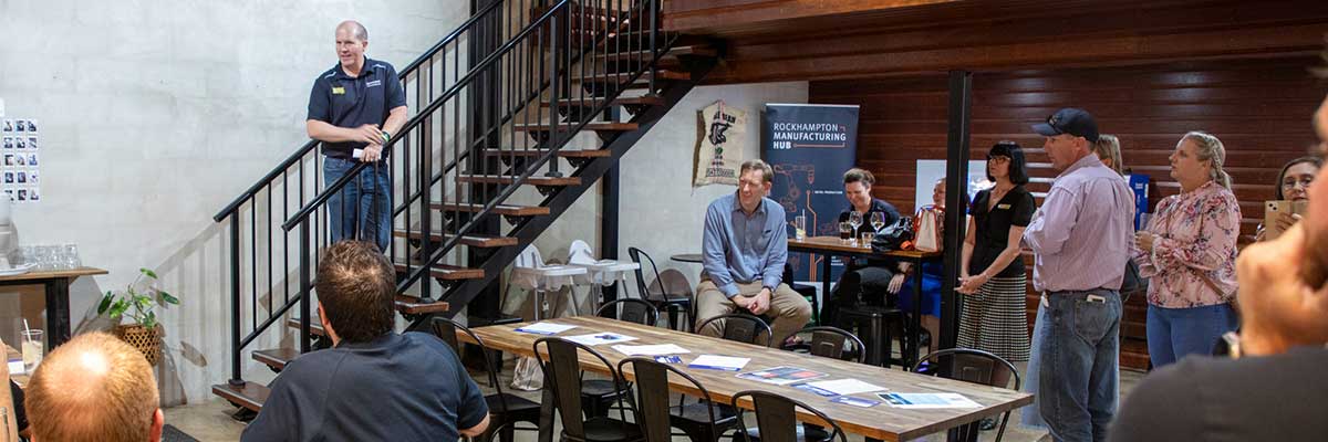 People sit and stand in a room, listening to a man deliver a presentation